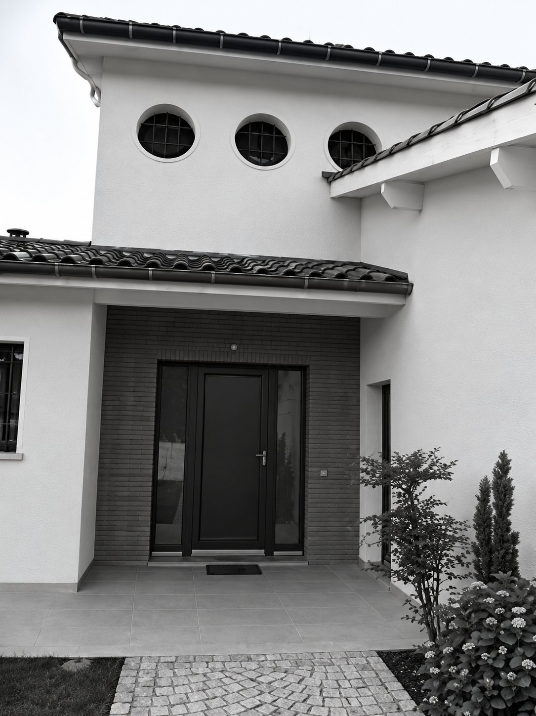 Photo en noir et blanc de la façade d’une maison contemporaine avec porte d’entrée sombre, trois petites fenêtres rondes et toit en tuiles.