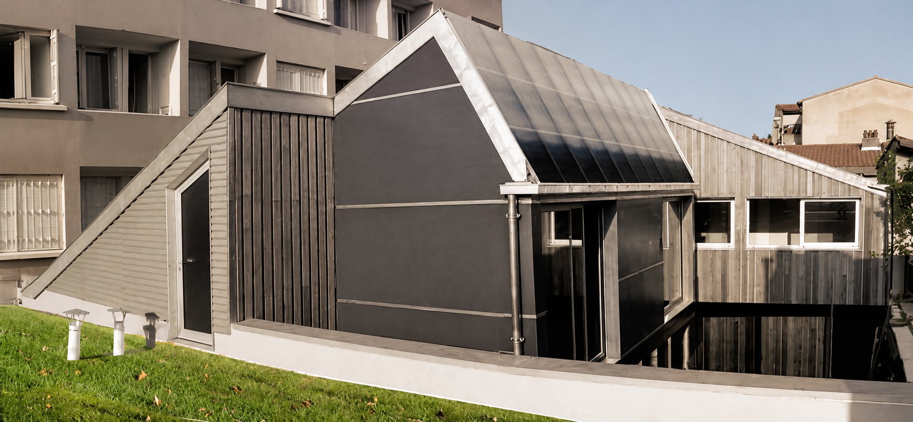 Extension d’architecture contemporaine avec bardage bois et toiture en zinc, construite près d’un immeuble, avec une pelouse au premier plan.