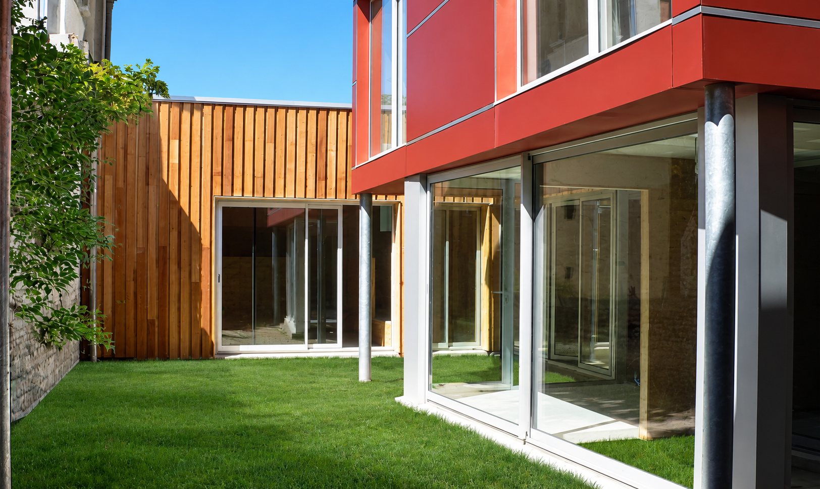 Cour engazonnée entourée d’une extension contemporaine avec bardage vertical en bois, façade rouge à l’étage et grandes baies vitrées coulissantes.