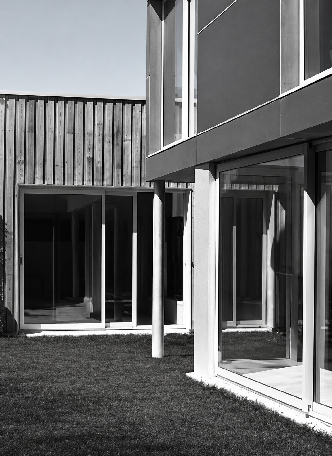 Photo en noir et blanc d’un bâtiment contemporain avec bardage en bois vertical, grandes baies vitrées et pelouse au premier plan.