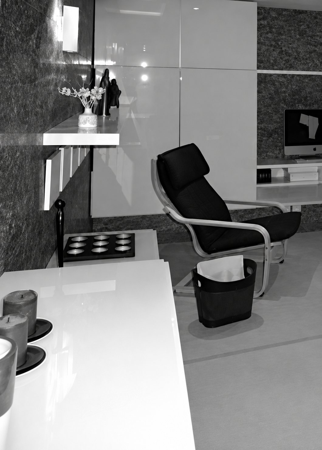 Photo en noir et blanc d’un intérieur moderne avec un fauteuil design, une table laquée blanche, une étagère murale et un écran au fond.