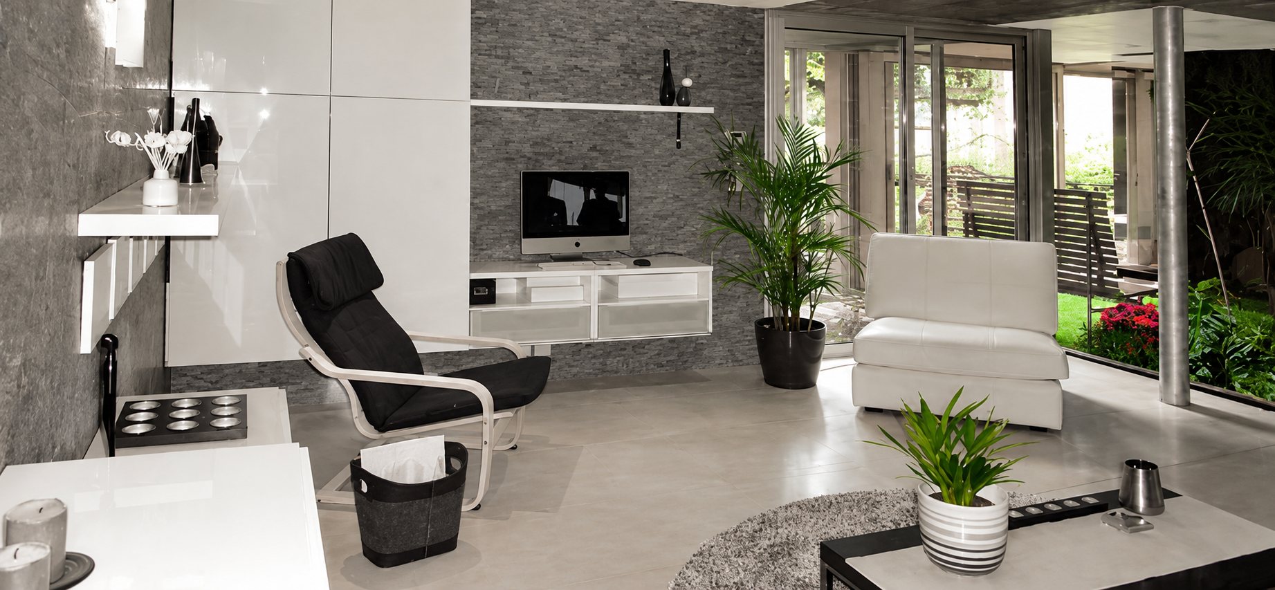 Salon contemporain aux tons gris et blancs avec fauteuil noir, canapé blanc, meuble télé suspendu, plantes vertes et grandes baies vitrées ouvrant sur un jardin.
