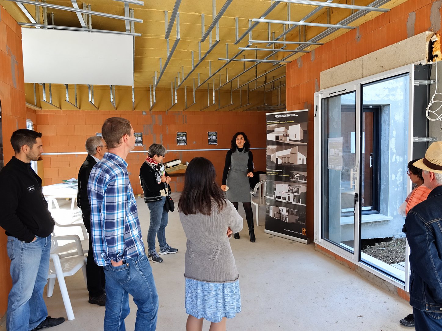 Un groupe de visiteurs écoute une femme qui présente une maison en construction aux murs en briques rouges, près d’une grande baie vitrée et d’une bannière « Prochains chantiers ».